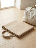 Sand Foldable Travel Yoga Mat folded flat beside a linen weekend bag